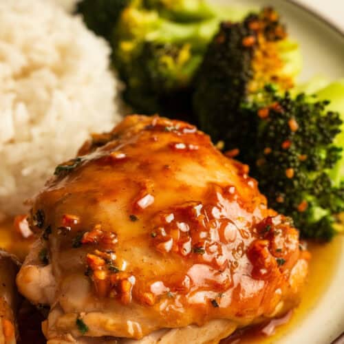 Plated crockpot honey garlic chicken thighs with a glossy honey garlic sauce, served with steamed white rice and roasted broccoli. The chicken is garnished with sesame seeds and chopped green onions, creating a vibrant and appetizing meal on a white ceramic plate.