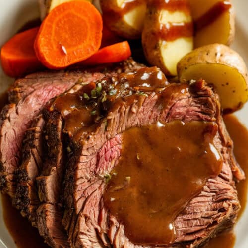 Plated slow cooker pot roast with tender beef, baby potatoes, and whole carrots, drizzled with a rich brown gravy. The hearty meal is served on a white ceramic plate, with warm lighting enhancing the rich colors and textures for an inviting presentation.