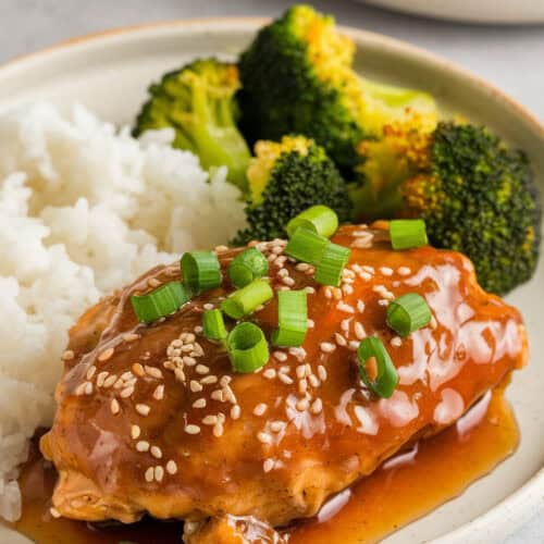 Slow cooker honey garlic chicken served with steamed white rice and roasted broccoli. The chicken is glazed in a thick honey garlic sauce, garnished with sesame seeds and green onions, creating a sweet and savory dish.