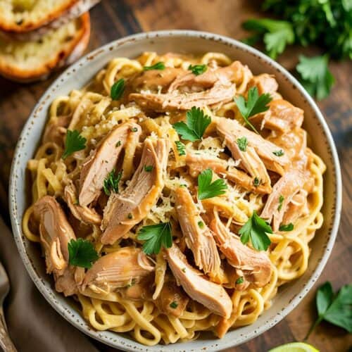 A photograph of a slow cooker chicken Marsala pasta served in a shallow white bowl, presented from a top-down perspective. The creamy, rich pasta is generously coated with shredded chicken and egg noodles, speckled with freshly grated Parmesan cheese and bright green parsley. The bowl sits invitingly on a warm, rustic wooden table alongside a golden-brown garlic bread and a few vibrant lime wedges, bathed in soft, natural light. The scene evokes a feeling of comfort and indulgence, showcasing a delicious homemade meal.