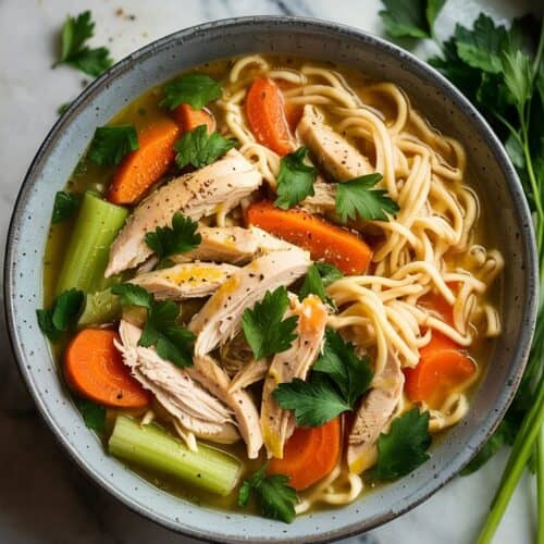 A photograph of a ceramic bowl brimming with a comforting chicken noodle soup viewed from directly above. Within the bowl are generous portions of shredded, golden-brown chicken, vibrant orange carrots, crisp green celery sticks, and perfectly cooked egg noodles suspended in a rich, savory broth. Freshly chopped parsley is sprinkled across the surface, alongside a delicate dusting of cracked black pepper, resting on a cool, polished marble kitchen countertop. Soft, natural light from a nearby window illuminates the scene, creating a warm and inviting ambiance.
