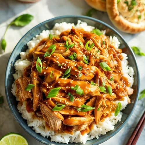A photograph of a top-down view showcasing slow cooker teriyaki chicken served over fluffy steamed rice on a pristine white marble kitchen countertop. The shredded chicken glistens under warm light, generously coated in a rich, creamy teriyaki sauce, and garnished with vibrant green onions and toasted sesame seeds. Arranged artfully around the rice are lime wedges and a golden-brown garlic breadstick, while soft morning light streams in from a nearby window, illuminating the scene. The composition creates an inviting and delectable presentation, highlighting the vibrant colors and textures of the dish.